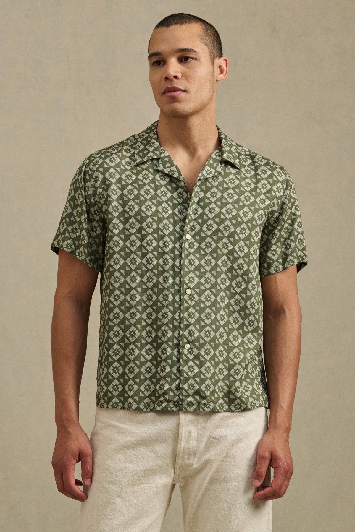 A man wears the EMILIO SHIRT by Velvet by Graham & Spencer—a green camp collar short-sleeve button-up with a white geometric pattern—and off-white pants, standing against a neutral background.-41479916355777