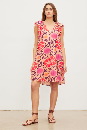 A woman wears the JENNA PRINTED SILK COTTON VOILE DRESS by Velvet by Graham & Spencer, paired with brown strappy sandals, posing against a plain white background. The v-neckline of the printed silk cotton voile fabric adds an elegant touch to her ensemble.