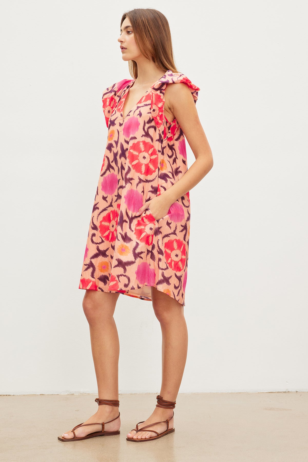 A woman stands sideways wearing a knee-length JENNA PRINTED SILK COTTON VOILE DRESS with flutter cap sleeves and a v-neckline by Velvet by Graham & Spencer. The printed silk cotton voile features a red and pink floral pattern. She is also wearing flat, brown sandals. She has long, straight hair. The background is plain white.-37073730568385