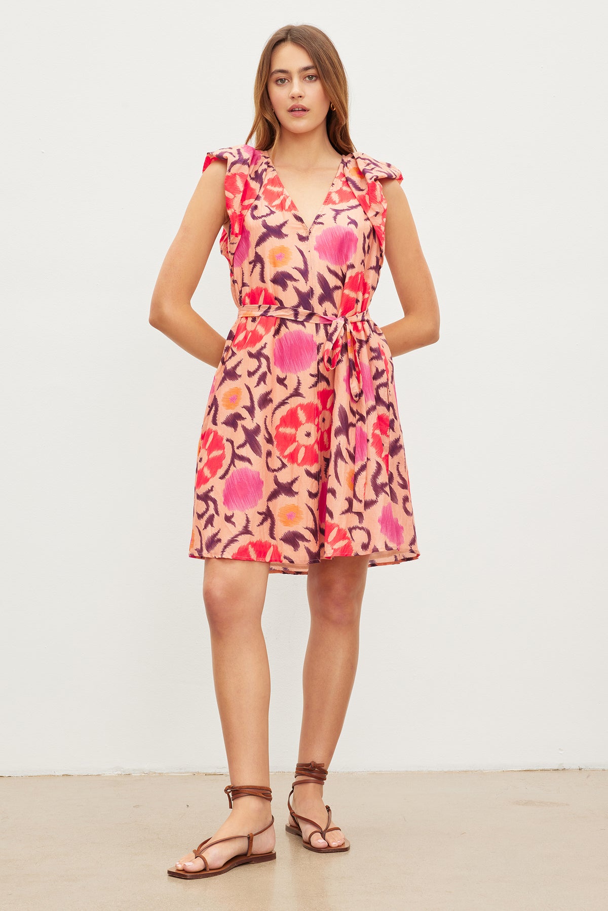 Woman standing against a plain background, wearing the Velvet by Graham & Spencer JENNA PRINTED SILK COTTON VOILE DRESS with a colorful floral pattern and flutter cap sleeves. She is also wearing brown sandals.