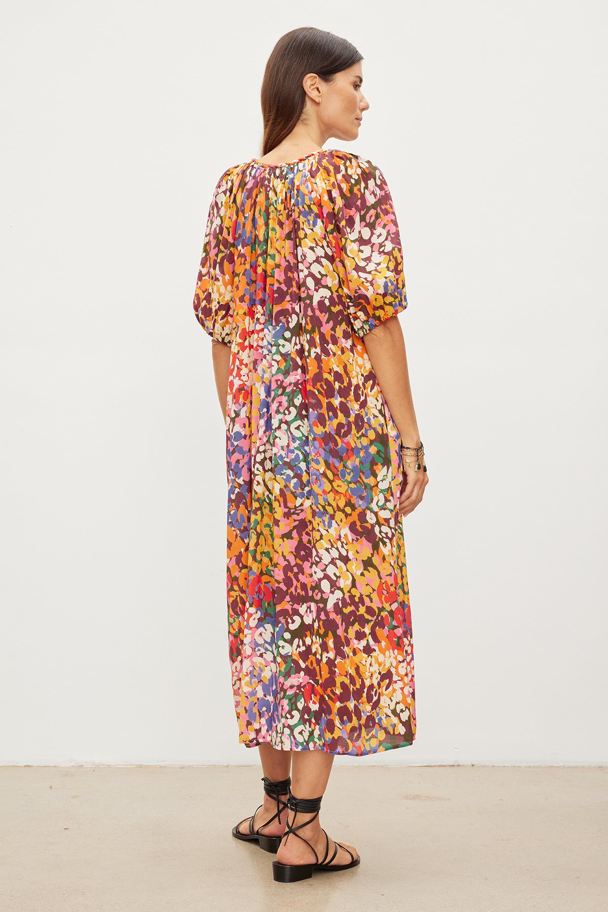 A woman viewed from the side wears a Velvet by Graham & Spencer CAROL PRINTED BOHO DRESS and black strappy sandals, standing against a plain white background.
