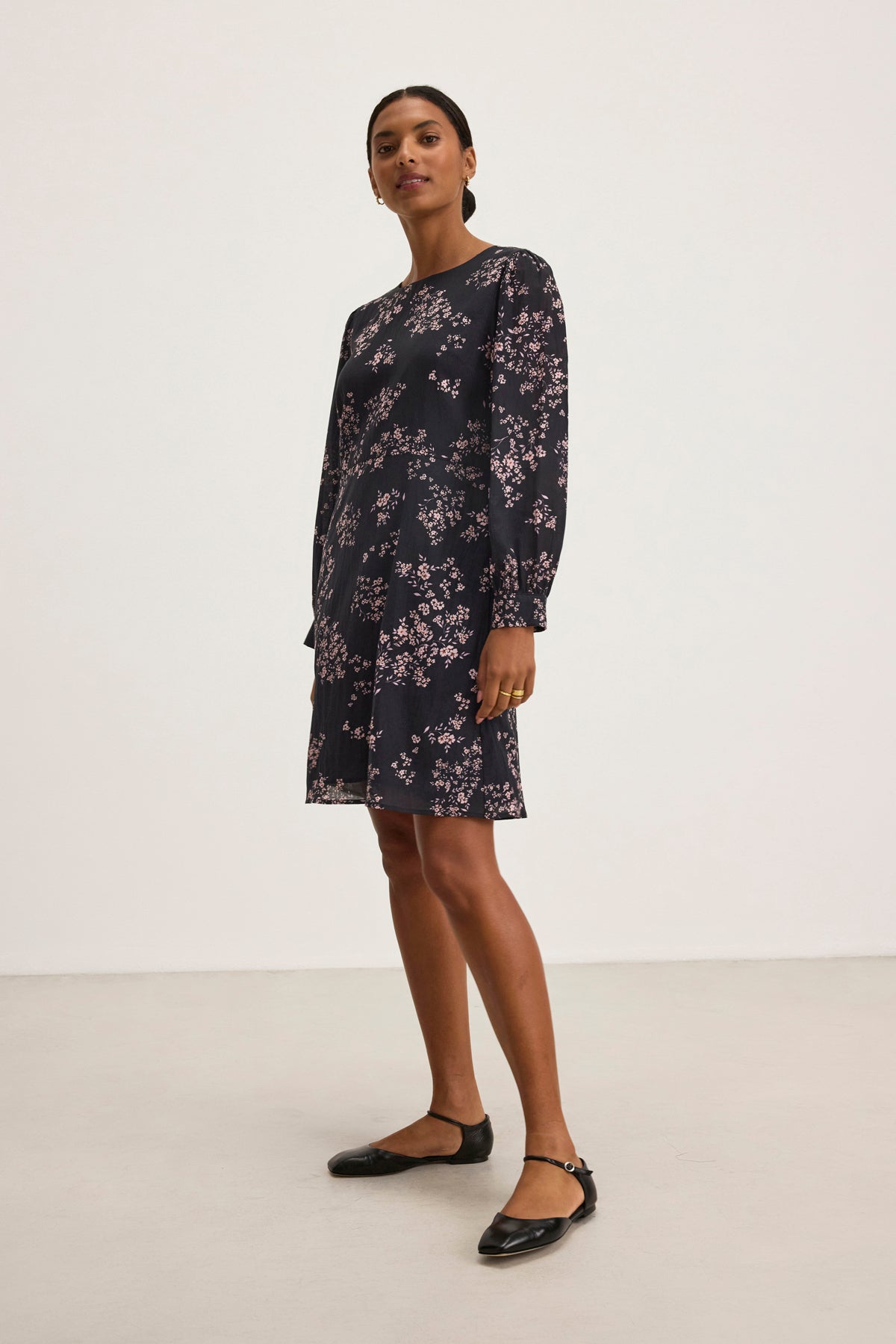 A person stands wearing the CELINA DRESS by Velvet by Graham & Spencer, featuring elegant small floral patterns in a cotton-viscose blend, paired with black flats, against a plain white background.
