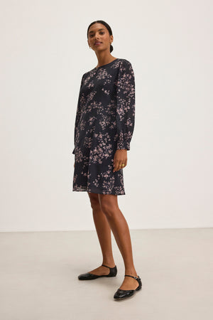 A person stands wearing the CELINA DRESS by Velvet by Graham & Spencer, featuring elegant small floral patterns in a cotton-viscose blend, paired with black flats, against a plain white background.