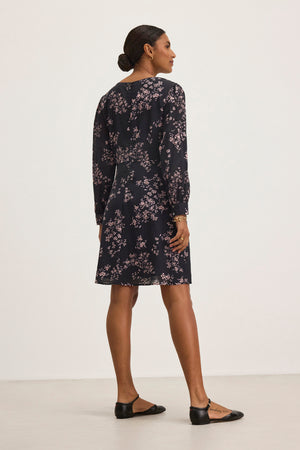 In a minimal setting, someone in the CELINA DRESS by Velvet by Graham & Spencer, an elegant dark floral cotton-viscose dress, pairs it with black flats while facing away.