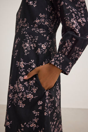 Close-up of someone in the elegant CELINA DRESS by Velvet by Graham & Spencer, made from cotton-viscose with pink floral patterns, pockets, and long sleeves. The breathable fabric ensures comfort as they casually place a hand in the pocket.