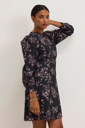 A woman in the CELINA DRESS by Velvet by Graham & Spencer, a black cotton-viscose piece with a pink floral pattern, stands against a plain white background, gazing at the camera with one hand on her head, exuding comfortable elegance.