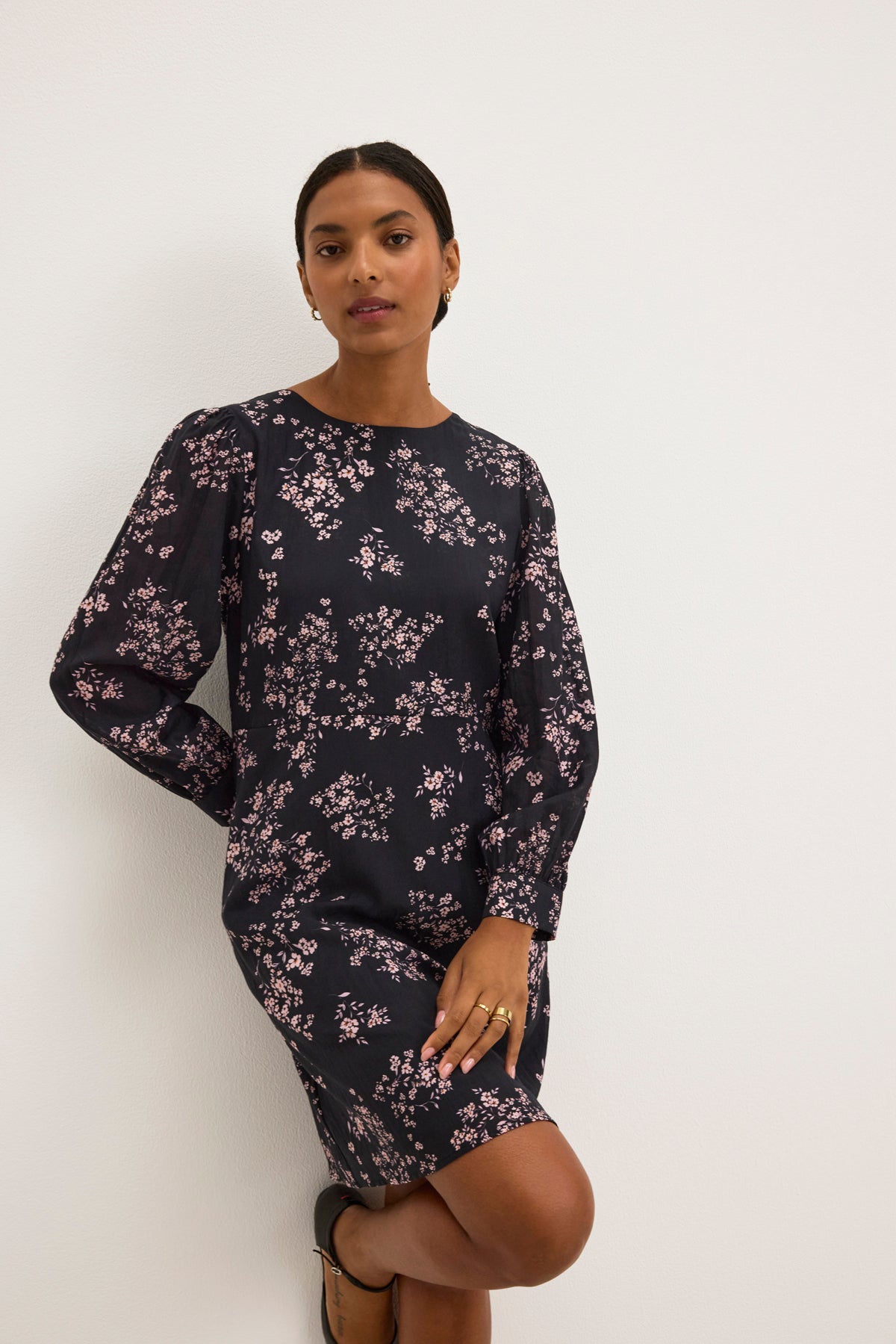 A person stands against a white wall wearing the elegant CELINA DRESS from Velvet by Graham & Spencer, featuring a dark floral design with pink accents, one hand on their hip and the other hanging down.-38943211159745