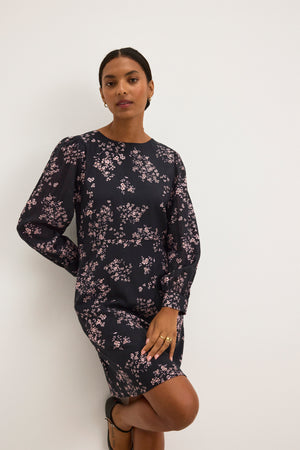 A person stands against a white wall wearing the elegant CELINA DRESS from Velvet by Graham & Spencer, featuring a dark floral design with pink accents, one hand on their hip and the other hanging down.