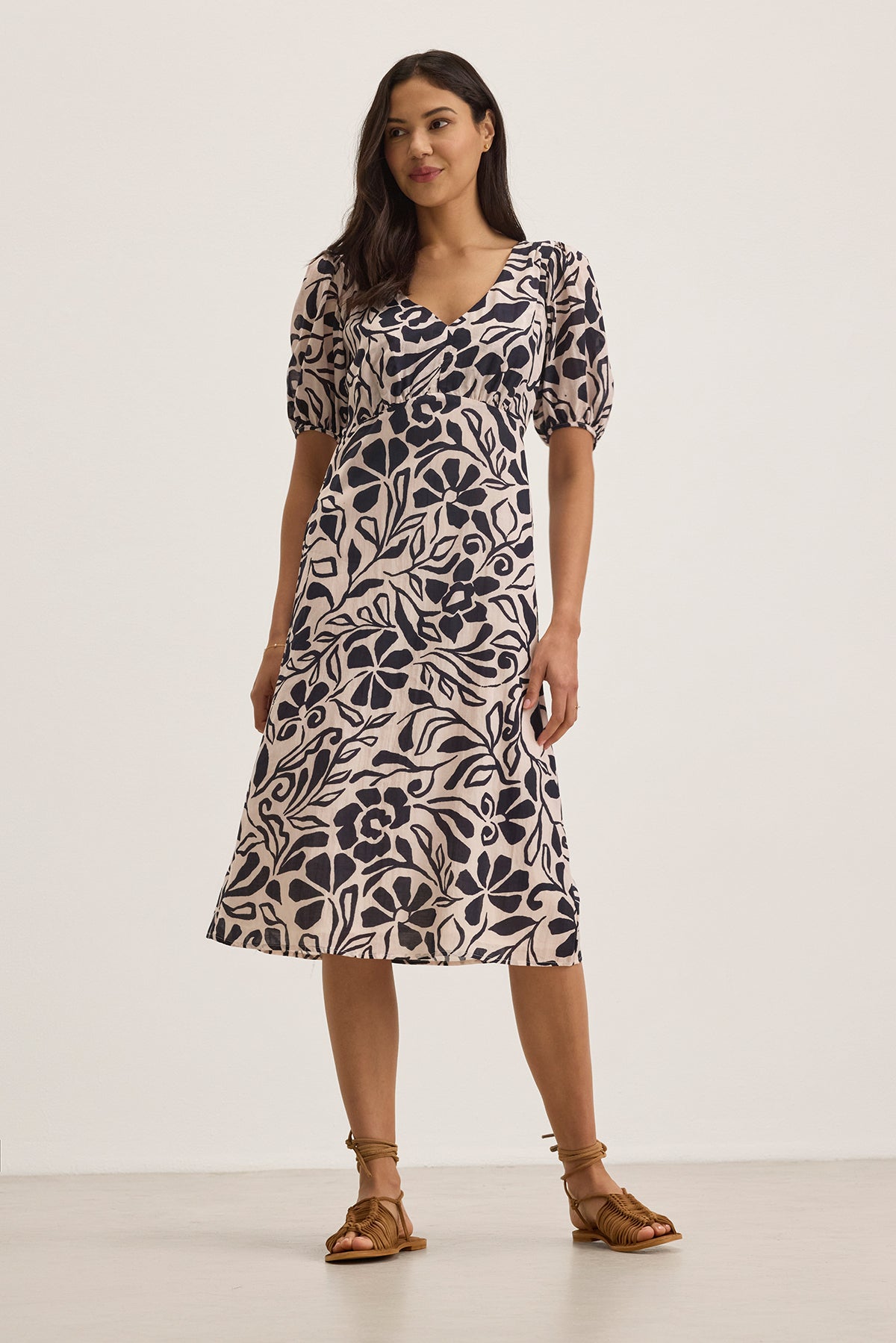 A woman poses in an INDIA DRESS by Velvet by Graham & Spencer, a cotton viscose midi dress adorned with black floral patterns on a pale background, featuring subtle puff sleeves.