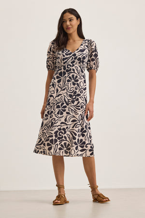 A woman poses in an INDIA DRESS by Velvet by Graham & Spencer, a cotton viscose midi dress adorned with black floral patterns on a pale background, featuring subtle puff sleeves.