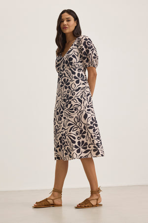 A woman in the INDIA DRESS by Velvet by Graham & Spencer, a floral-patterned cotton viscose midi with puff sleeves, pairs it with flat sandals against a plain background.