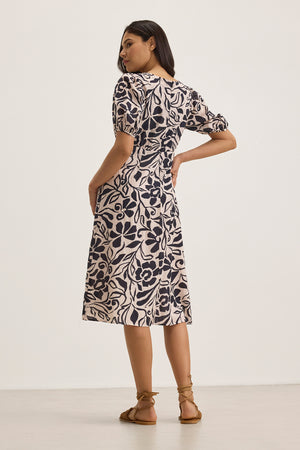 A woman stands on a light floor, facing away, wearing the INDIA DRESS by Velvet by Graham & Spencer—a knee-length, floral-patterned cotton viscose midi dress with puff sleeves.
