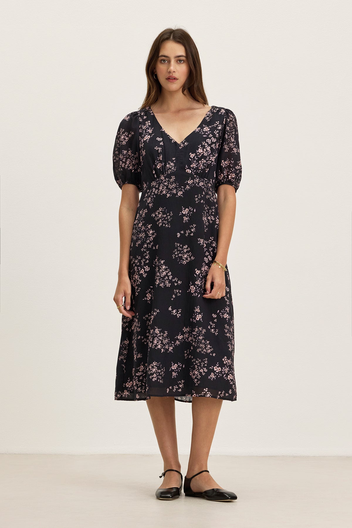 A woman stands against a plain background wearing the INDIA DRESS by Velvet by Graham & Spencer—a black, floral-patterned midi dress with short sleeves—paired with black flat shoes.-39333209604289