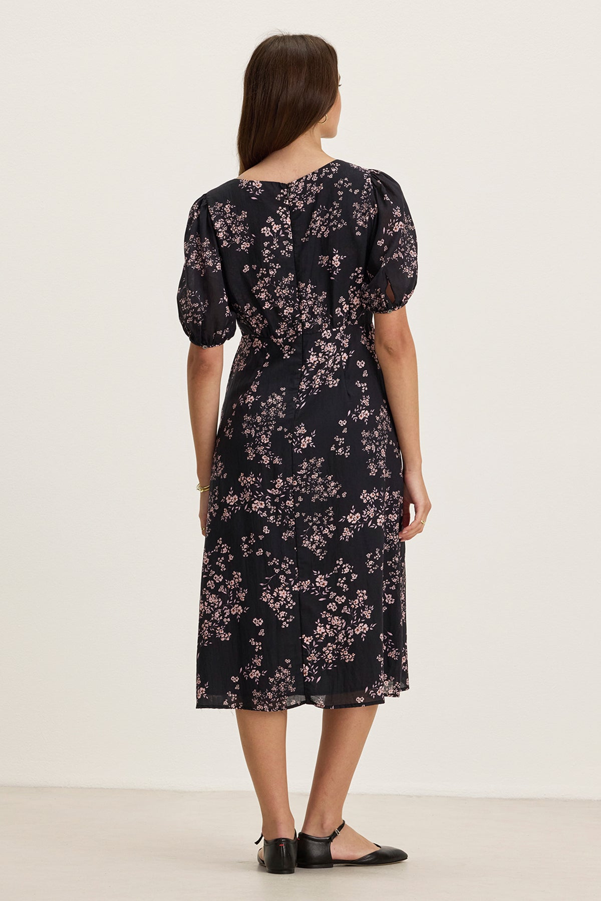 Woman standing indoors, facing away, wearing the INDIA DRESS by Velvet by Graham & Spencer—a black midi dress with pink floral pattern, v-neckline, short puff sleeves—and black flat shoes.-39333209669825