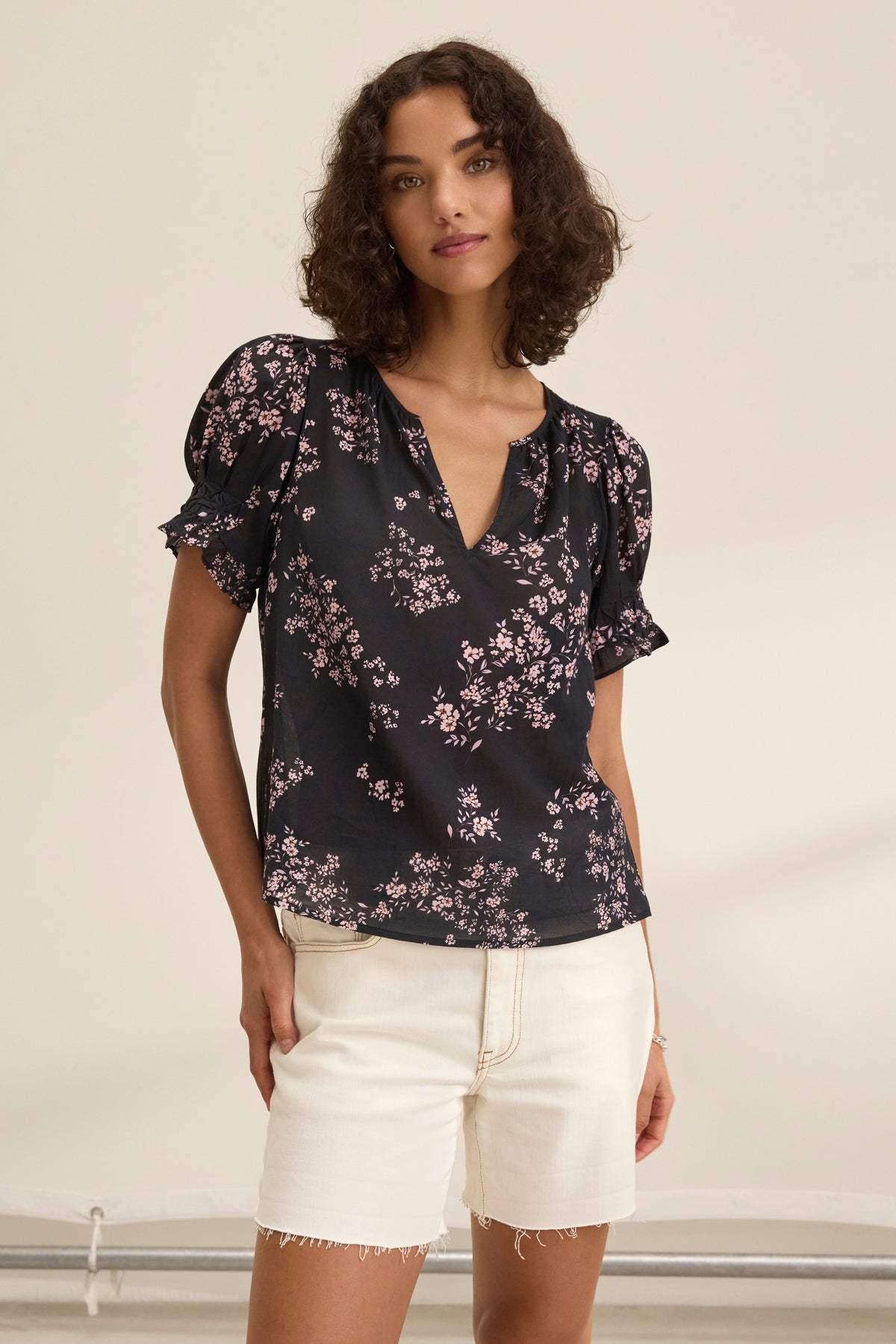 Someone with curly hair is wearing the Velvet by Graham & Spencer LESLIE TOP, a black floral blouse with puff sleeves and a relaxed fit, paired with white shorts against a plain background.-38943211061441