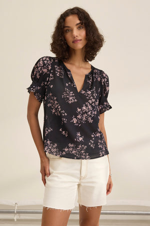 Someone with curly hair is wearing the Velvet by Graham & Spencer LESLIE TOP, a black floral blouse with puff sleeves and a relaxed fit, paired with white shorts against a plain background.
