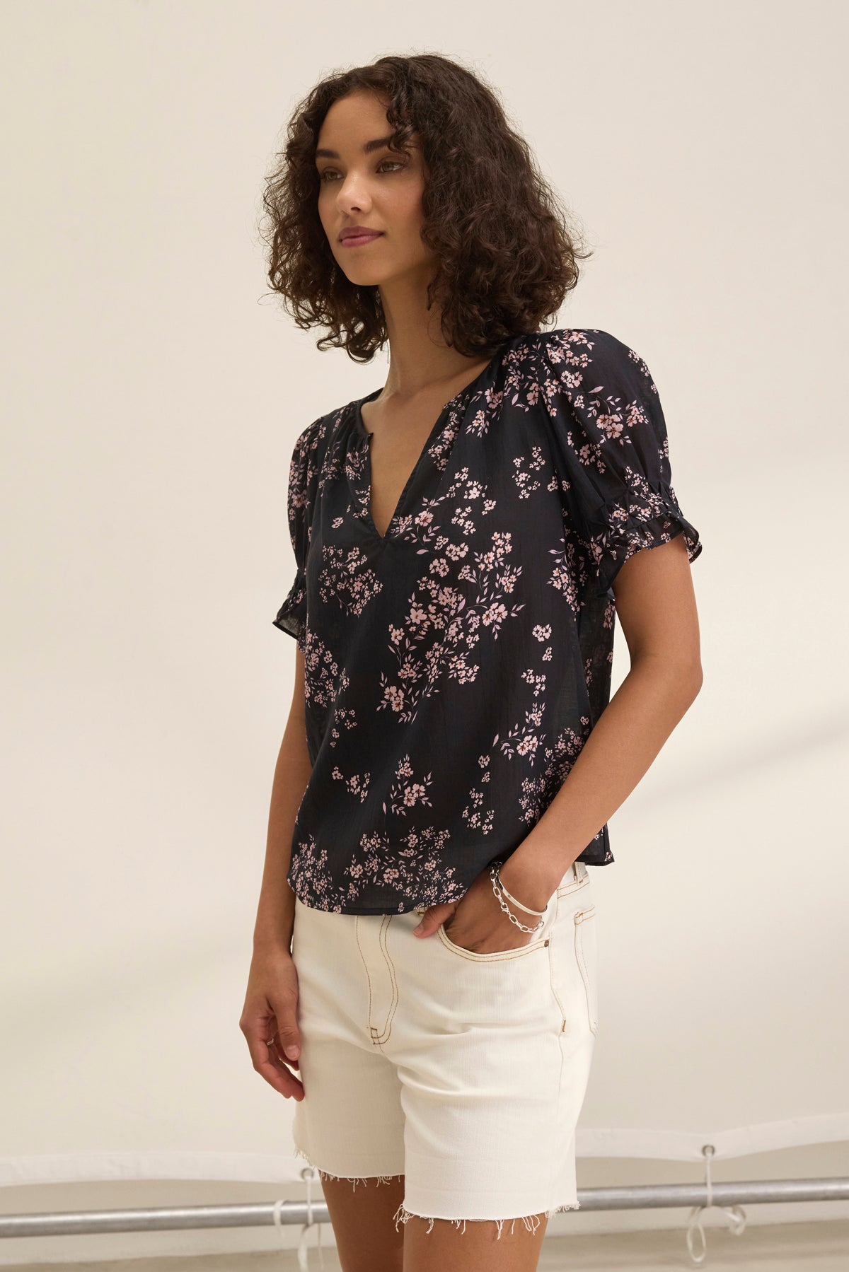 A woman with curly hair is wearing the LESLIE TOP, a black floral blouse by Velvet by Graham & Spencer, featuring puff sleeves and a relaxed fit. She pairs it with white shorts against a plain background.-38943211028673
