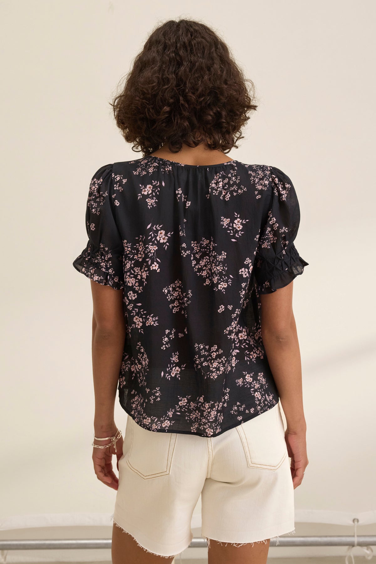 A person with curly hair is wearing the LESLIE TOP, a black floral blouse featuring a relaxed fit and V-neckline by Velvet by Graham & Spencer, paired with light shorts. They stand with their back to the camera.-38943211094209