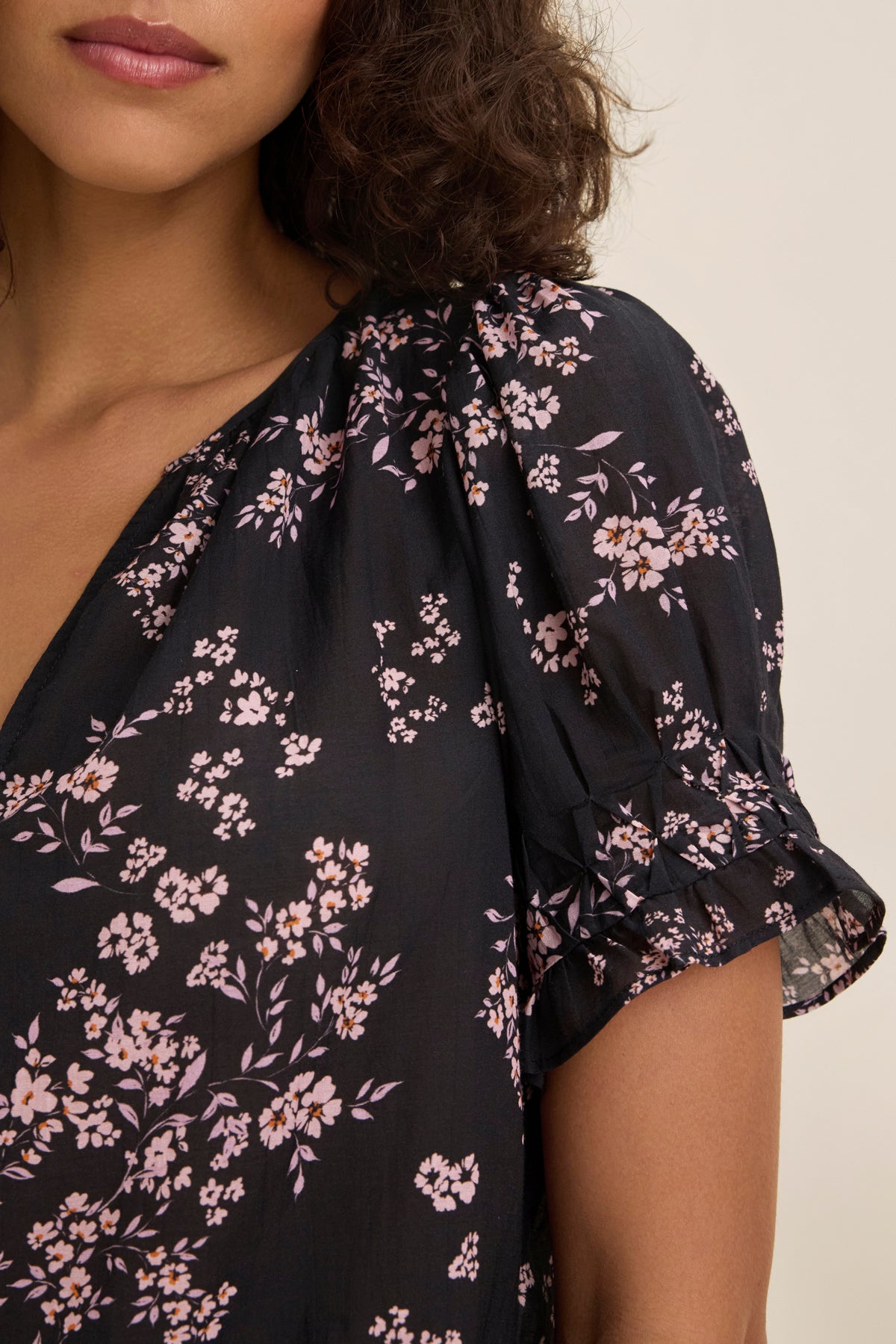 A woman models the LESLIE TOP by Velvet by Graham & Spencer, showcasing a black design with pink floral patterns, short puff sleeves, and a V-neckline against a plain backdrop.