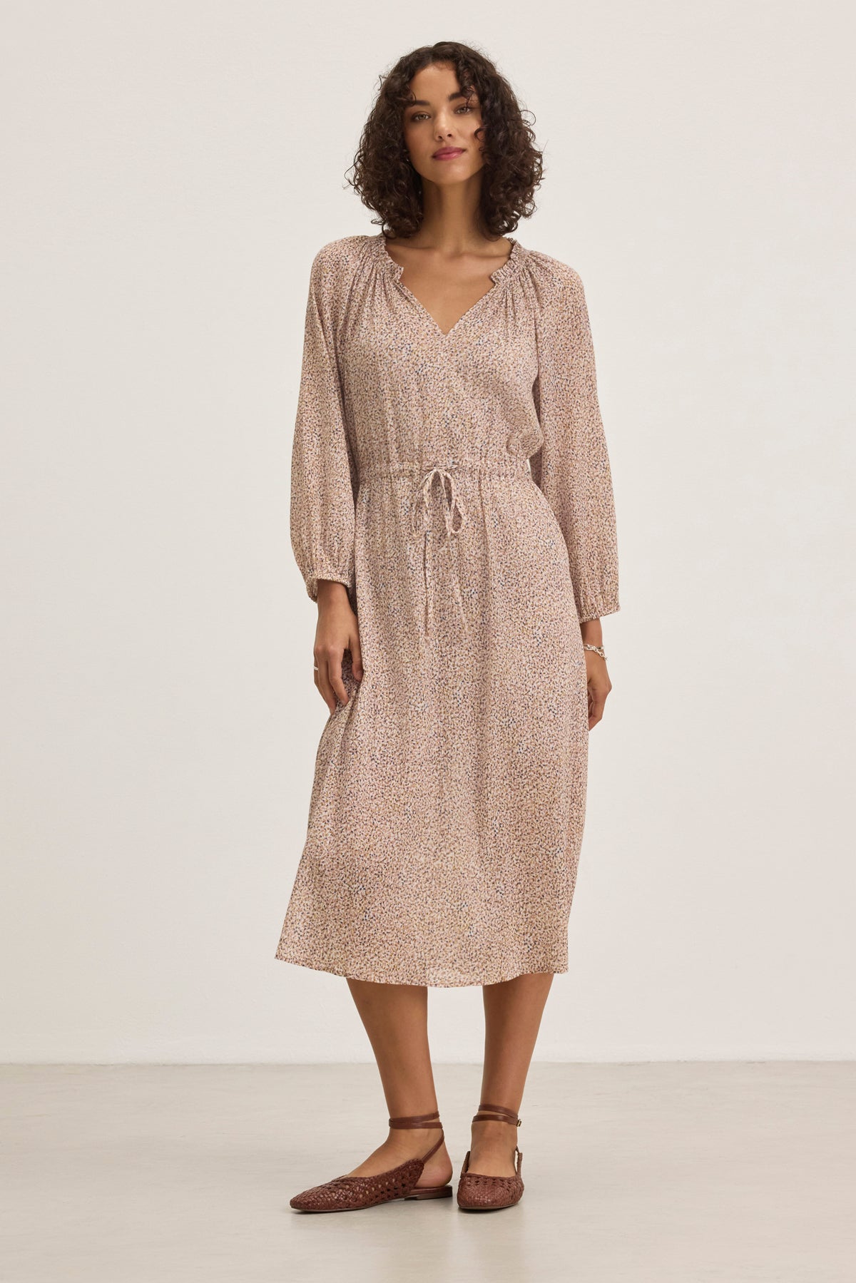 A woman in a Velvet by Graham & Spencer LIMA PRINTED CRINKLE COTTON GAUZE DRESS—a light brown, long-sleeved, knee-length piece with subtle patterns—stands against a plain background. She's wearing brown flat shoes and has curly hair.-38820676403393