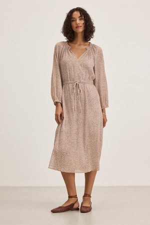 A woman in a Velvet by Graham & Spencer LIMA PRINTED CRINKLE COTTON GAUZE DRESS—a light brown, long-sleeved, knee-length piece with subtle patterns—stands against a plain background. She's wearing brown flat shoes and has curly hair.
