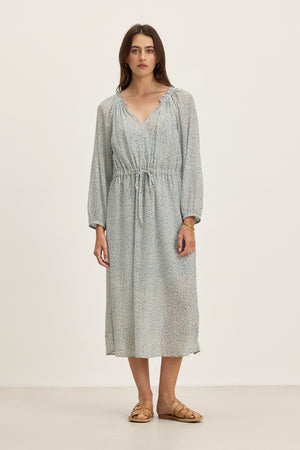 A woman stands against a plain background wearing the LIMA PRINTED CRINKLE COTTON GAUZE DRESS by Velvet by Graham & Spencer, featuring a ruffled V-neckline, drawstring waist, and tan sandals.