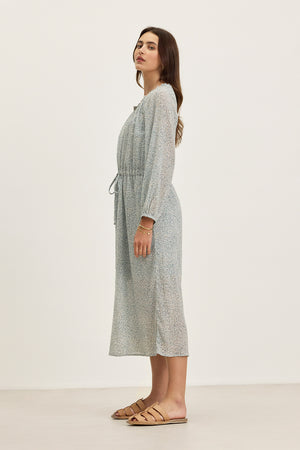 A woman stands sideways wearing the LIMA PRINTED CRINKLE COTTON GAUZE DRESS by Velvet by Graham & Spencer, featuring a light blue print, ruffled V-neckline, waist tie, and tan flat sandals against a plain background.