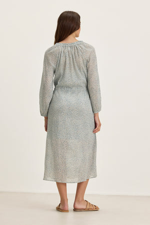 A woman faces away in the Velvet by Graham & Spencer LIMA PRINTED CRINKLE COTTON GAUZE DRESS—a light blue, patterned midi with a ruffled V-neckline—paired with tan flat sandals against a plain light background.
