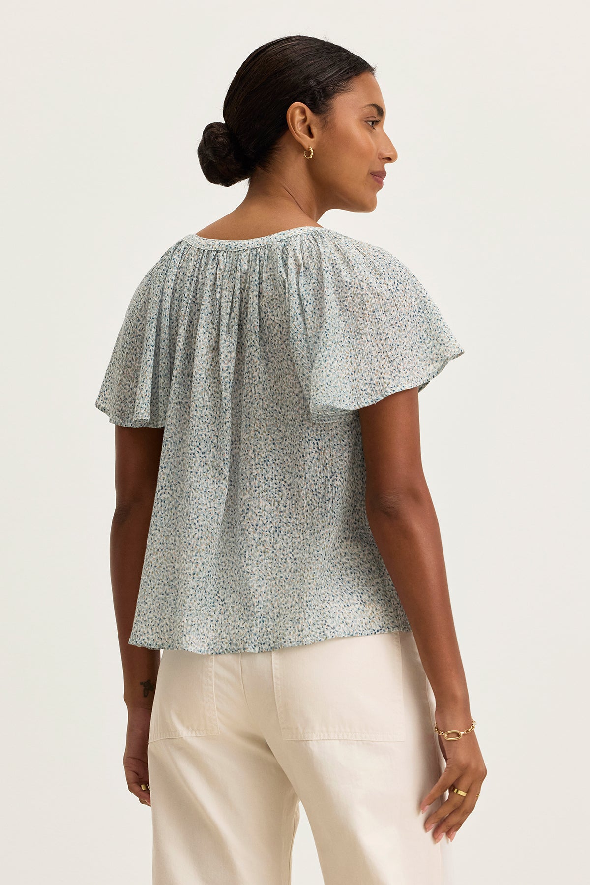 A woman with her hair in a low bun stands facing away, wearing the Velvet by Graham & Spencer PARK PRINTED CRINKLE COTTON GAUZE TOP in light blue with white pants.-39333214716097