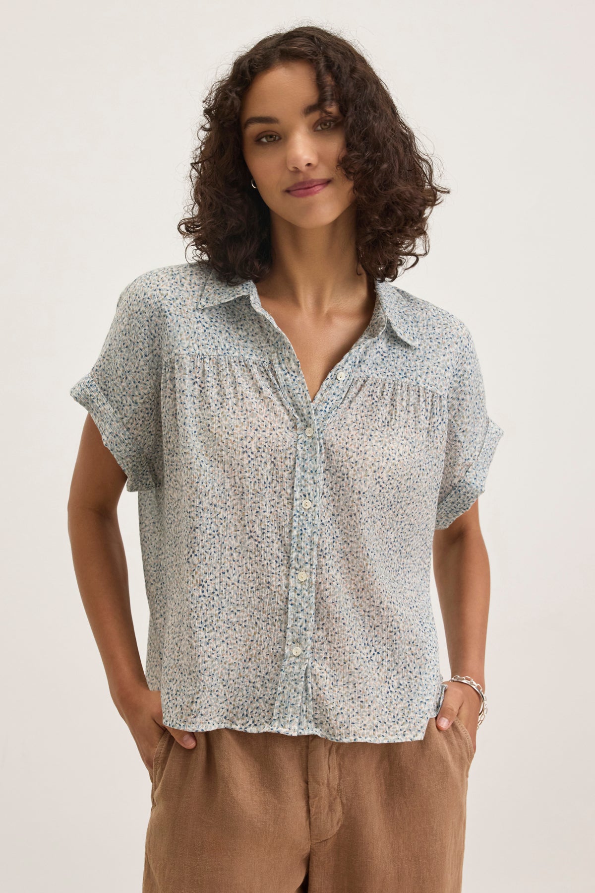 A person with curly hair stands against a plain background, wearing the TIGE PRINTED CRINKLE COTTON GAUZE TOP by Velvet by Graham & Spencer in light blue and relaxed fit, paired with tan pants.