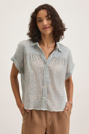 A person with curly hair stands against a plain background, wearing the TIGE PRINTED CRINKLE COTTON GAUZE TOP by Velvet by Graham & Spencer in light blue and relaxed fit, paired with tan pants.