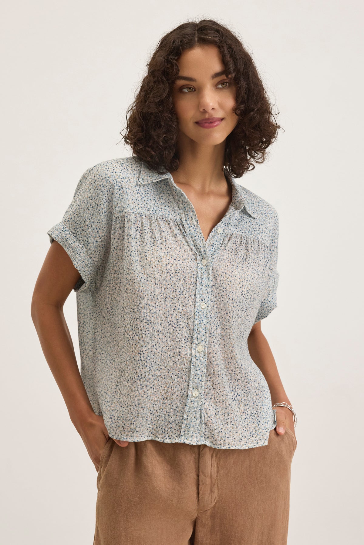 A woman with curly hair wears the TIGE PRINTED CRINKLE COTTON GAUZE TOP by Velvet by Graham & Spencer in light blue and beige pants, standing against a plain background.-38957722763457