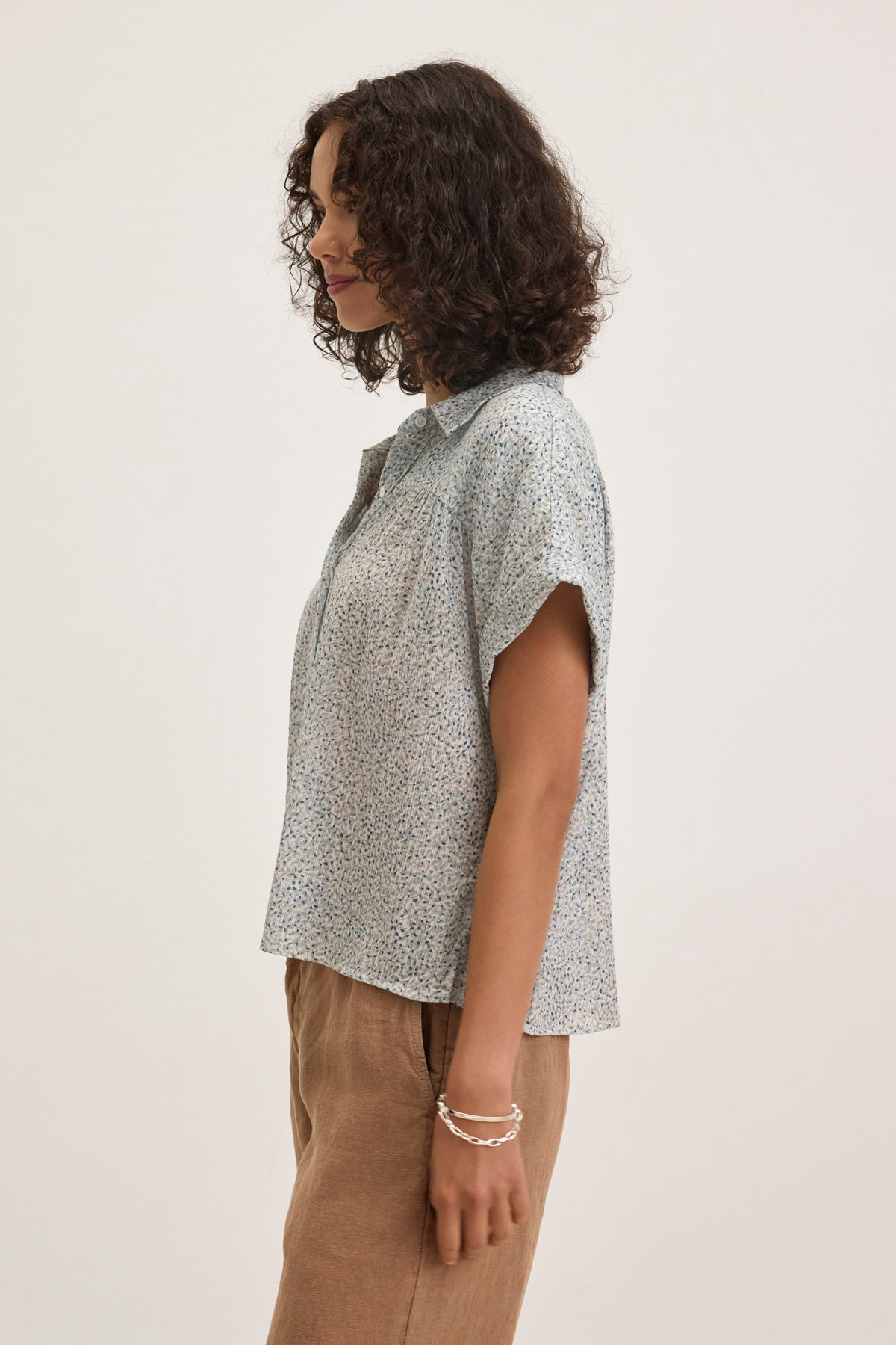A person with curly hair stands in profile wearing the TIGE PRINTED CRINKLE COTTON GAUZE TOP by Velvet by Graham & Spencer, paired with light brown pants against a plain background.-38957722730689