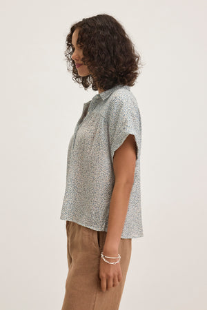 A person with curly hair stands in profile wearing the TIGE PRINTED CRINKLE COTTON GAUZE TOP by Velvet by Graham & Spencer, paired with light brown pants against a plain background.