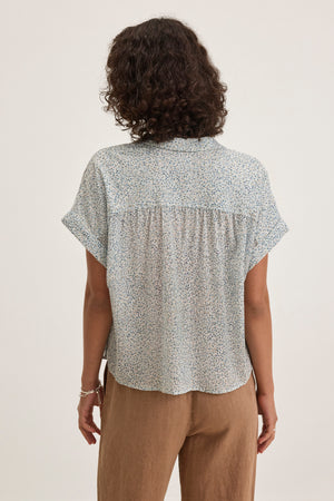 A person with curly hair is seen from the back, wearing a Velvet by Graham & Spencer TIGE PRINTED CRINKLE COTTON GAUZE TOP and brown pants, standing against a plain background.