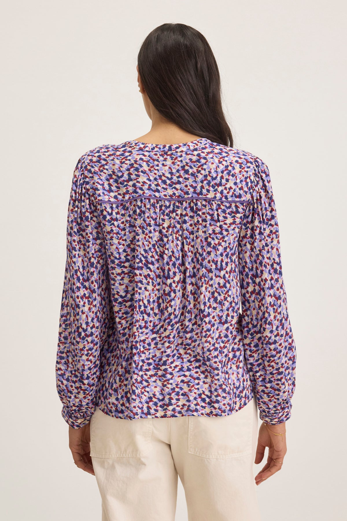 A person with long dark hair faces away, wearing the ELDA TOP by Velvet by Graham & Spencer, a colorful and patterned blouse, paired with white pants against a plain background.-38749660807361