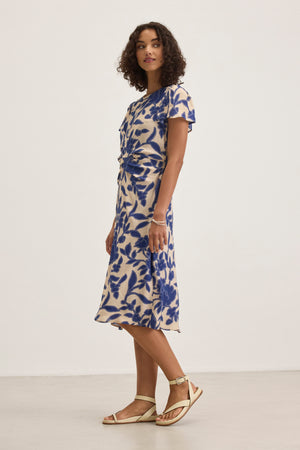 A person wearing the NEVINE DRESS by Velvet by Graham & Spencer, featuring a blue and white floral pattern, stands against a plain background.