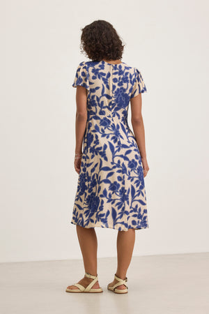 A person is seen from the back, wearing the NEVINE DRESS with a blue floral pattern, by Velvet by Graham & Spencer, paired with beige sandals against a plain light background.
