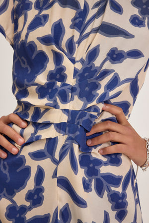 A close-up features a person wearing the NEVINE DRESS by Velvet by Graham & Spencer. The beige dress boasts blue floral patterns, with hands on the waist and a silver bracelet adorning their right wrist.