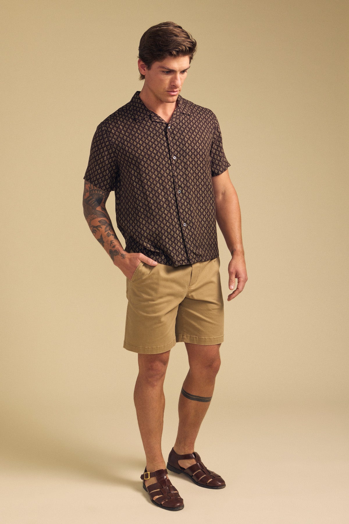A man stands against a beige background, wearing the EMILIO SHIRT by Velvet by Graham & Spencer—a relaxed fit, patterned camp collar shirt in breathable modal—paired with tan shorts, brown sandals, and showing tattoos on his left arm and leg.-41701960024257