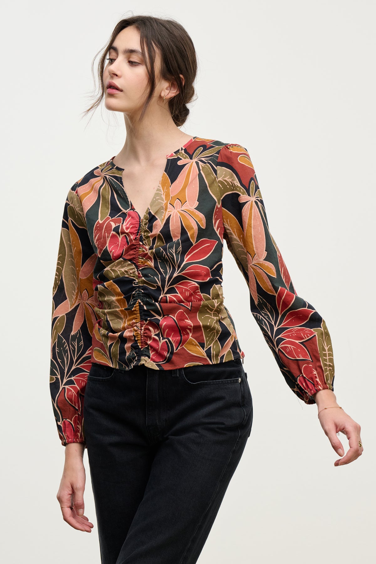 A woman wears the SYDNEY TOP by Velvet by Graham & Spencer, featuring long sleeves, a V-neckline, colorful leaf print, and ruched details, paired with black jeans against a plain light background.-39742395678913