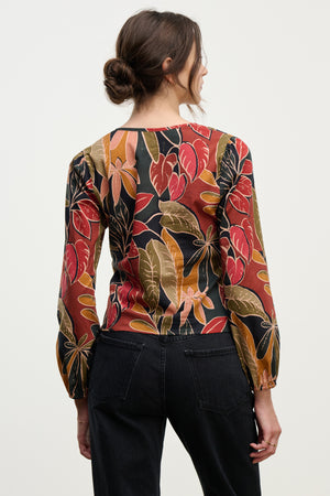 A woman with brown hair in a low bun, wearing the SYDNEY TOP by Velvet by Graham & Spencer—a long-sleeved printed cotton cambric blouse with a colorful leaf pattern—stands facing away from the camera.