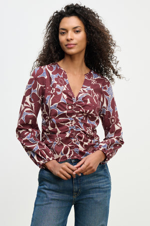 A woman wears the SYDNEY TOP by Velvet by Graham & Spencer—a long-sleeve, ruched, floral cotton cambric blouse in burgundy tones with blue and white patterns—paired with blue jeans, standing against a plain light background.