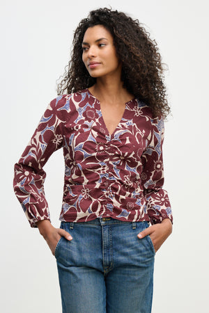 A woman wears the SYDNEY TOP by Velvet by Graham & Spencer, a long-sleeve floral maroon V-neck blouse with blue and white accents, paired with blue jeans, standing with hands in pockets against a plain background.