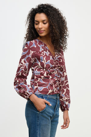 A woman with curly hair wears the SYDNEY TOP by Velvet by Graham & Spencer—a maroon, printed cotton cambric blouse with a V-neckline—paired with blue jeans, standing against a plain background with one hand in her pocket.