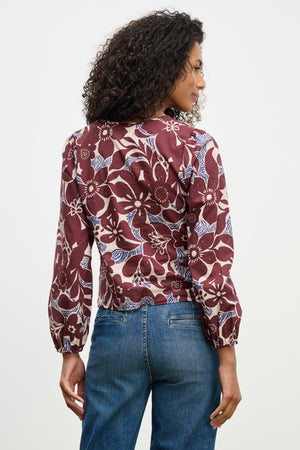 A woman with curly hair is seen from the back, wearing the SYDNEY TOP by Velvet by Graham & Spencer—a long-sleeved, V-neck floral blouse in maroon and blue printed cotton cambric—paired with blue jeans.