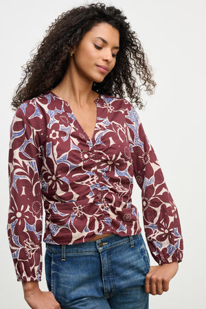 A woman wears the SYDNEY TOP by Velvet by Graham & Spencer—a maroon and blue floral cotton cambric long-sleeve, ruched V-neck blouse—paired with blue jeans, standing against a plain light background.