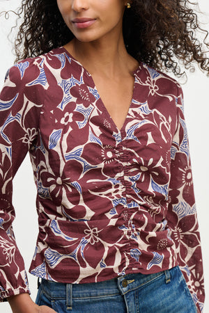 A person wears the SYDNEY TOP by Velvet by Graham & Spencer—a maroon floral-printed cotton cambric blouse with a V-neckline and blue, white accents—paired with blue jeans against a plain background.