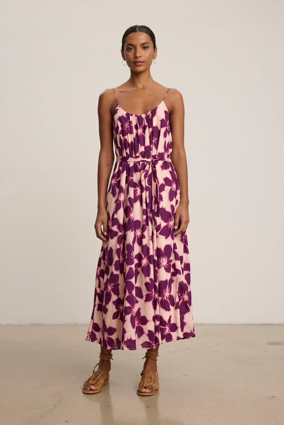 A woman wears the ALEXA DRESS by Velvet by Graham & Spencer—a sleeveless, ankle-length cotton voile maxi dress with a purple floral print on light pink, finished with a matching belt and styled with brown sandals.-39111850623169