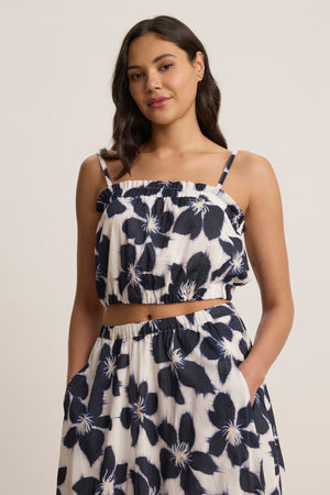 A woman wears the ALICE CAMI by Velvet by Graham & Spencer—an adjustable-strap, cropped floral cami paired with a high-waisted, black and white voile skirt, standing with her hands in the skirt’s pockets.
